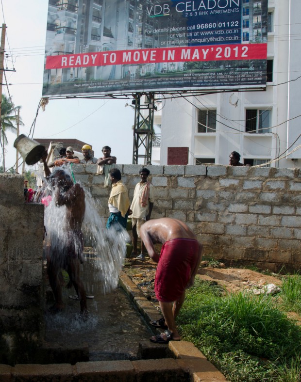 Construction workers taking a bucket bath in front of a billboard marketing a middle-class gated community in Bangalore - by SabinavonKessel - wikimedia commons